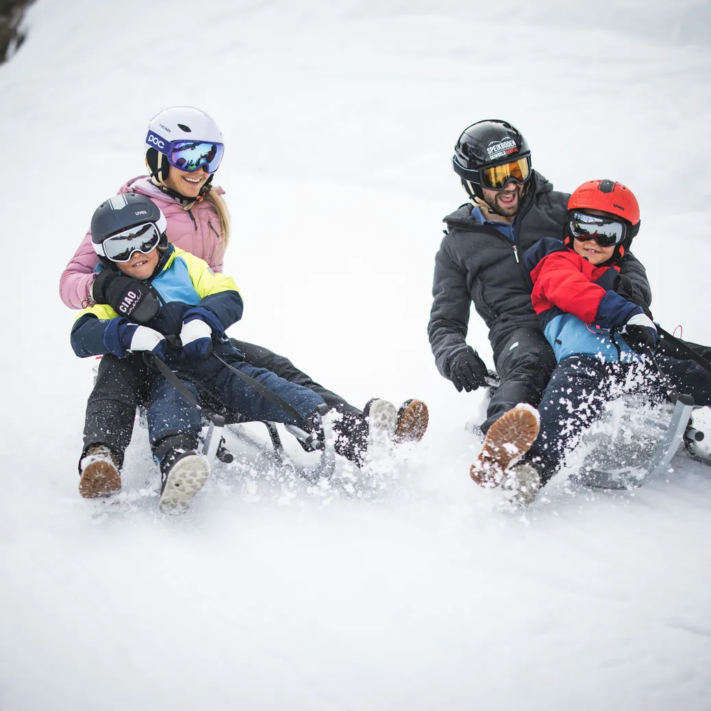 Family sledging on the toboggan run – parents and children with helmets on snowy slope