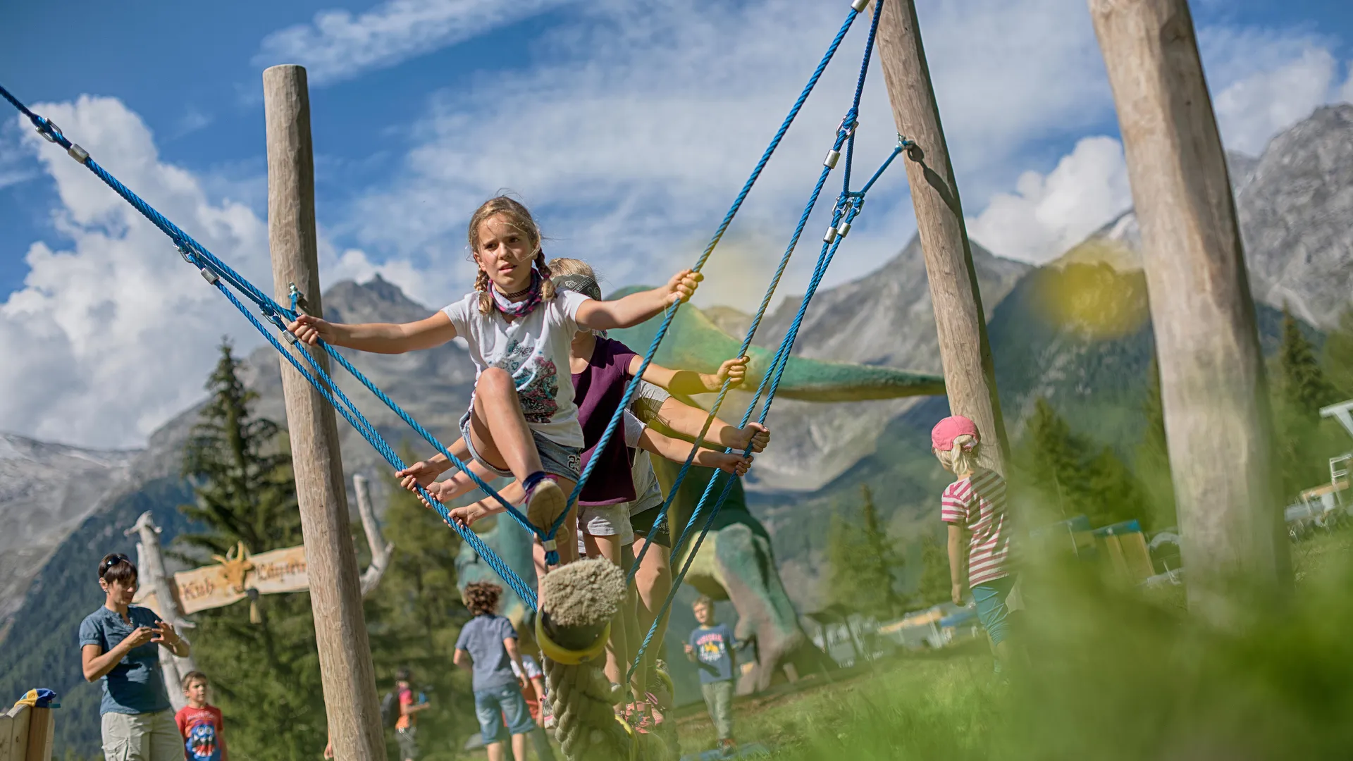 Kinder spielen auf großer Seilschaukel am Erlebnispark mit Dinosaurier-Figur und Bergpanorama Klausberg