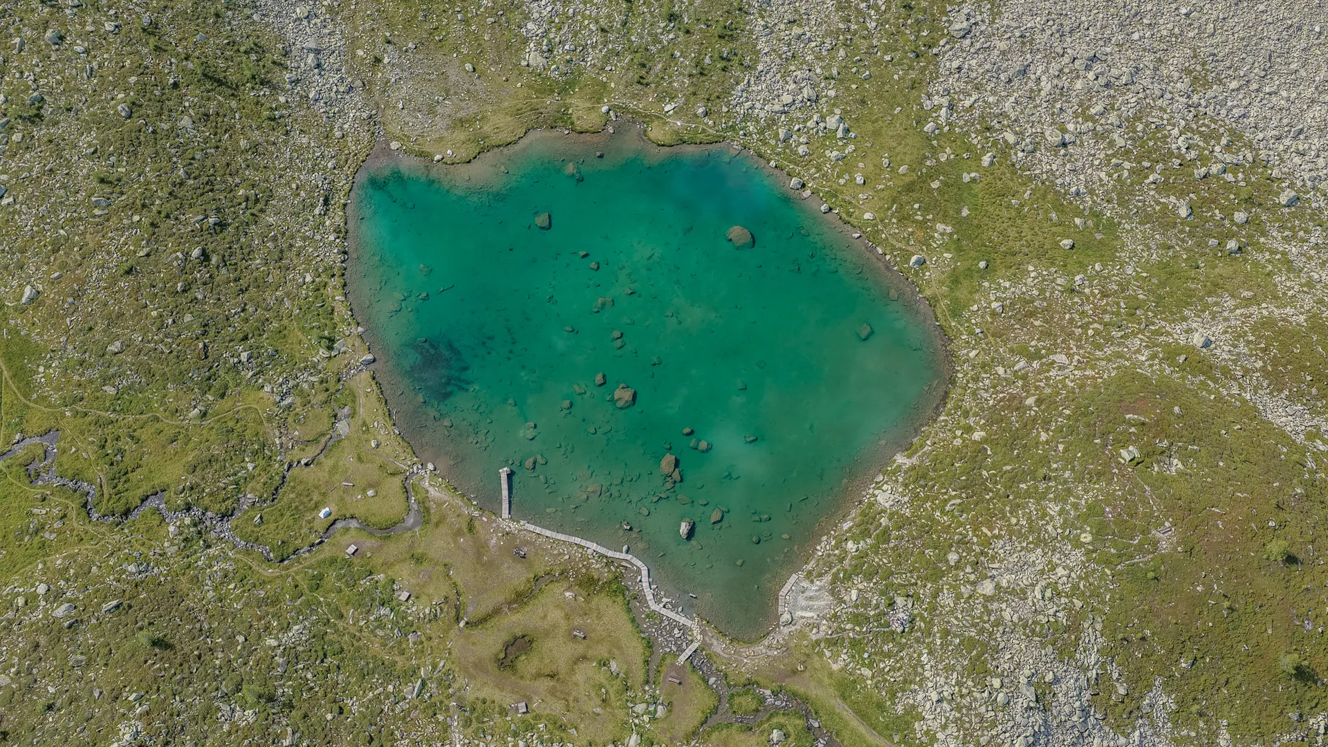 Luftaufnahme des türkisfarbenen Bergsees am Speikboden mit Holzsteg, Geröll und Almwiesen