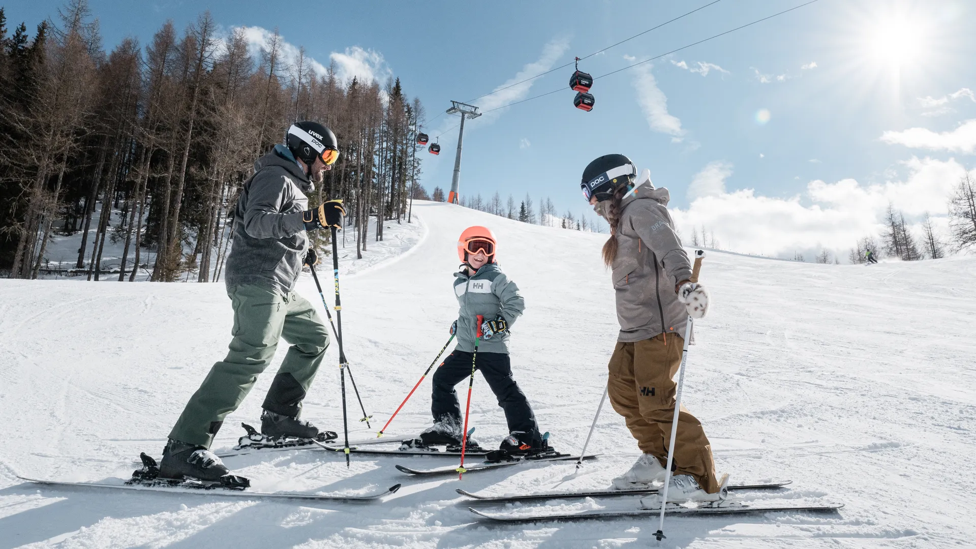 Family skiing on a sunny slope at Klausberg with red cable car gondolas in the background