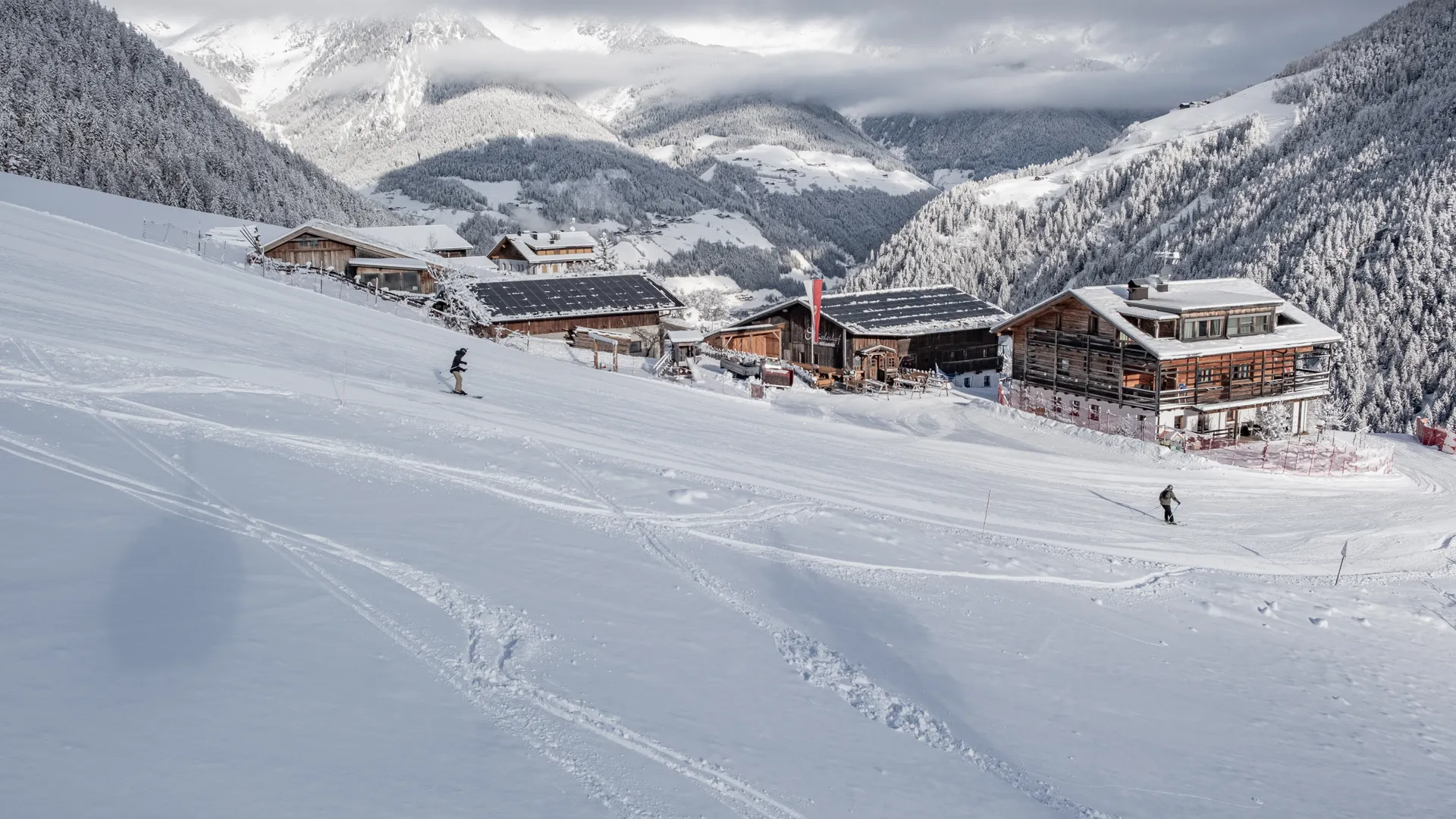 Snowboarder and skier on Klausberg run with alpine huts and snowy Ahrntal valley panorama