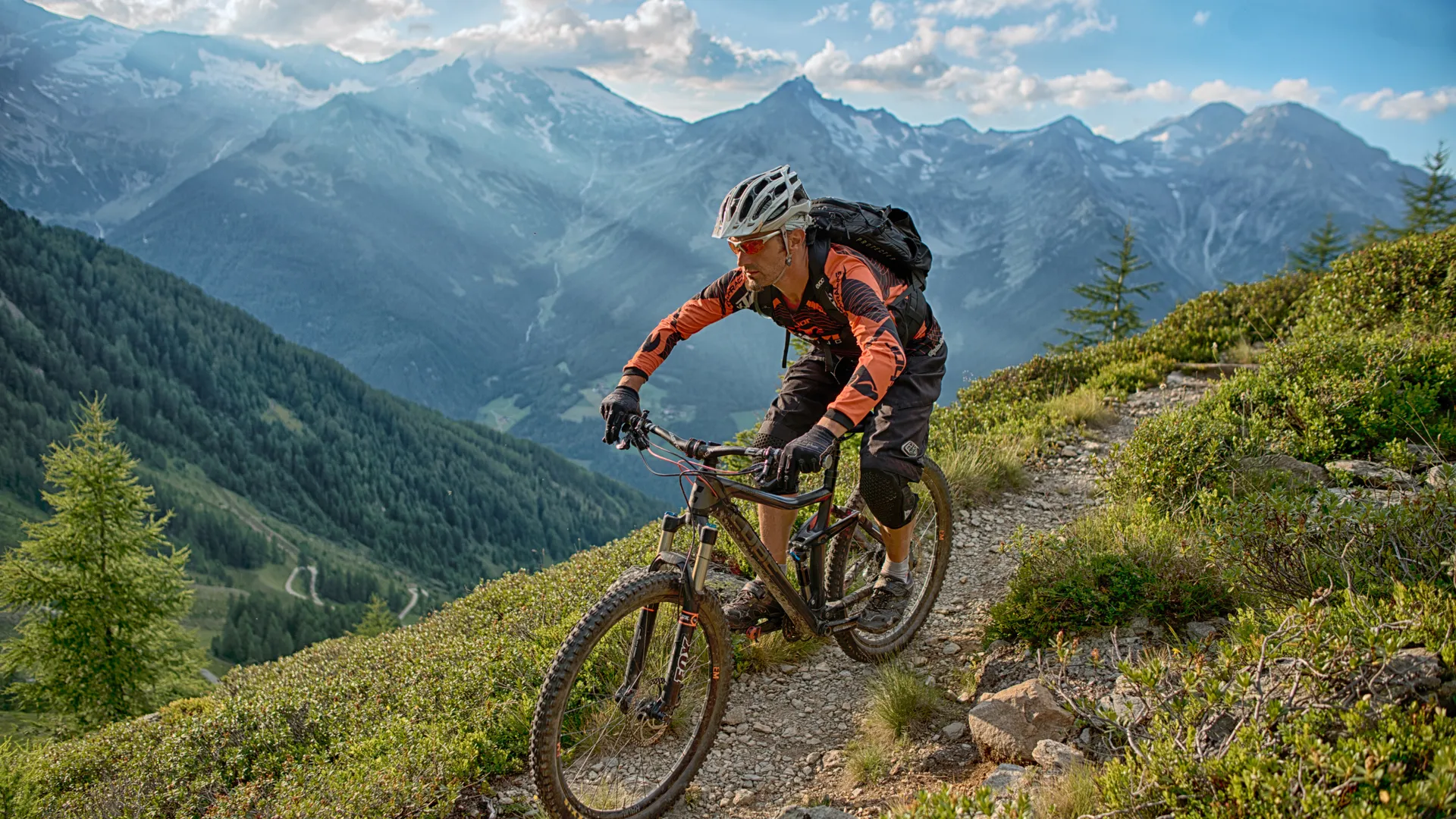 Mountainbiker in oranger Kleidung fährt auf Singletrail durch Almlandschaft mit Panorama der Zillertaler Alpen