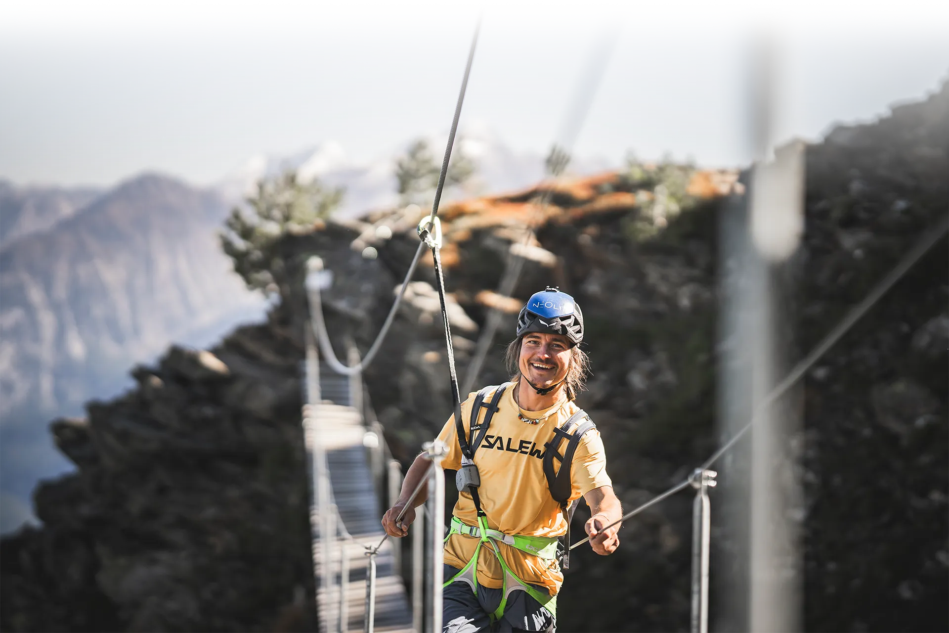 Lachender Klettersteig-Geher mit blauem Helm auf Hängebrücke, Bergpanorama Ahrntal