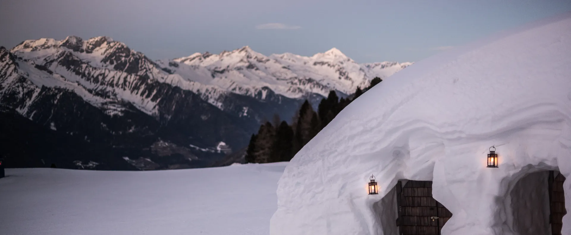 Illuminated igloo with wooden door and lanterns at dusk with Alpine panorama at Klausberg