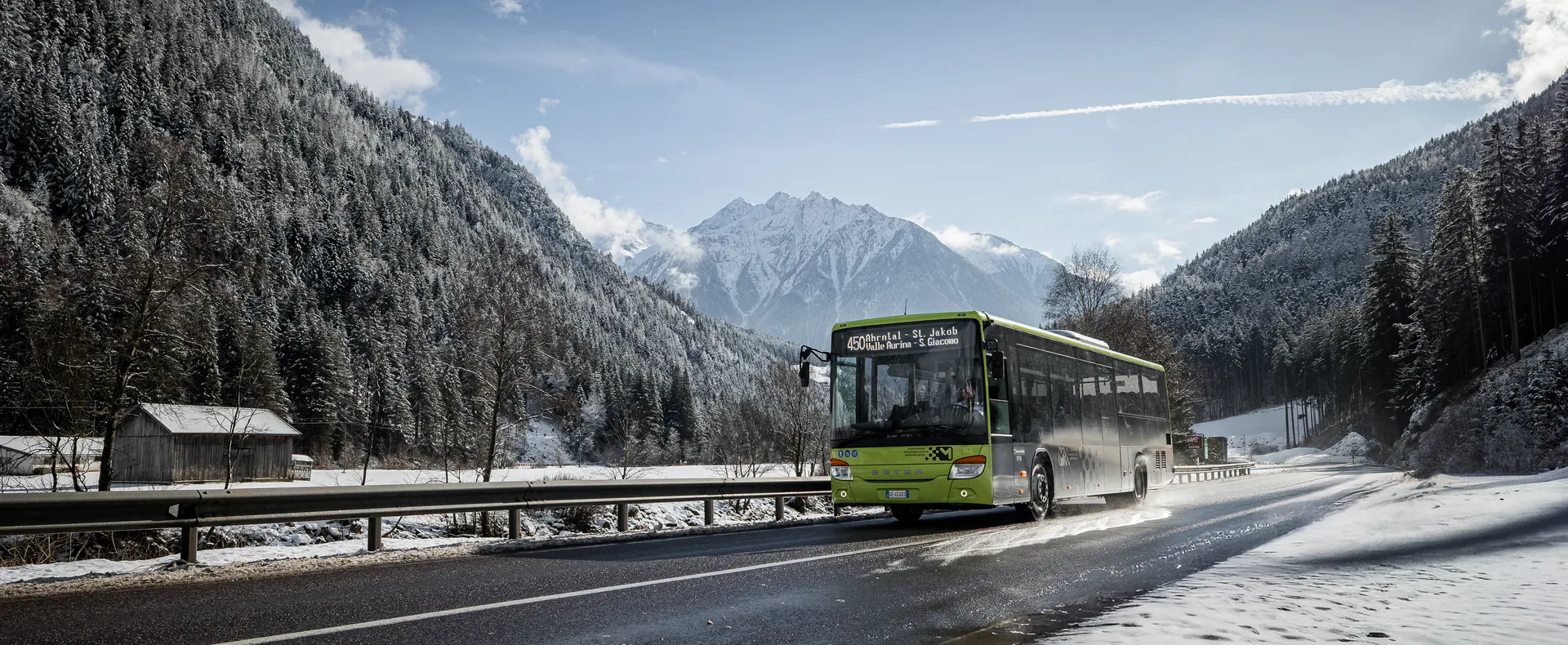Green ski bus on Ahrntal line drives through snowy valley with forest and mountains