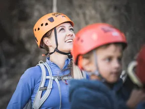 Junge Frau mit orangem Speikboden-Kletterhelm lächelt strahlend, Kind mit rotem Helm davor