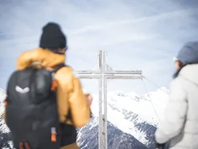 Hölzernes Gipfelkreuz am Speikboden mit Schneeschuhwanderern und weitem Alpenpanorama im Ahrntal