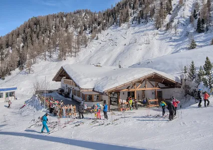Sonnige Skihütte mit Gästen auf der Terrasse und Skiständern, bewaldete Berghänge
