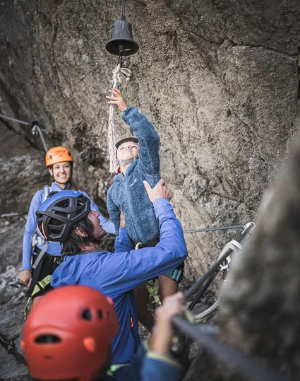 Family on Ahrntal via ferrata: child rings summit bell, parents with helmets celebrate together
