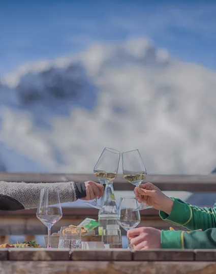 Paar stößt mit Weißwein auf Hüttenterrasse an, schneebedeckte Berge im Hintergrund