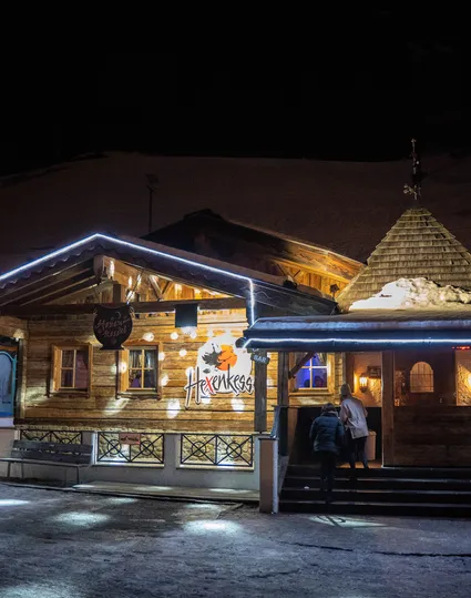 Hexenkessel Après-Ski-Bar bei Nacht beleuchtet, Schnee und Gondelbahn im Hintergrund