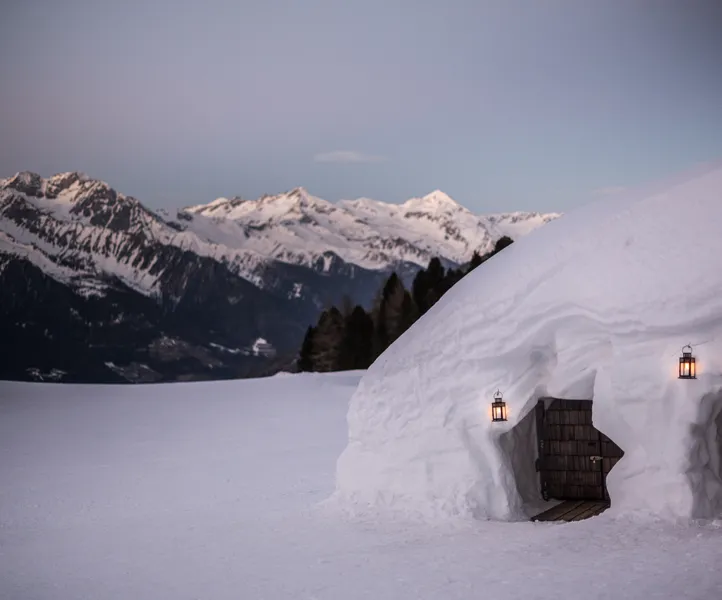 Beleuchtetes Iglu mit Holztür und Laternen in der Abenddämmerung mit Alpenpanorama am Klausberg