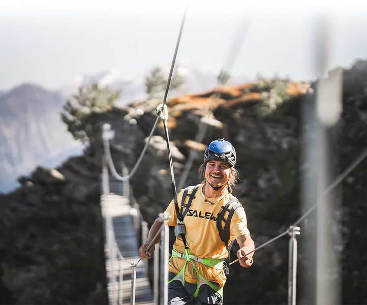 Lachender Klettersteig-Geher mit blauem Helm auf Hängebrücke, Bergpanorama Ahrntal
