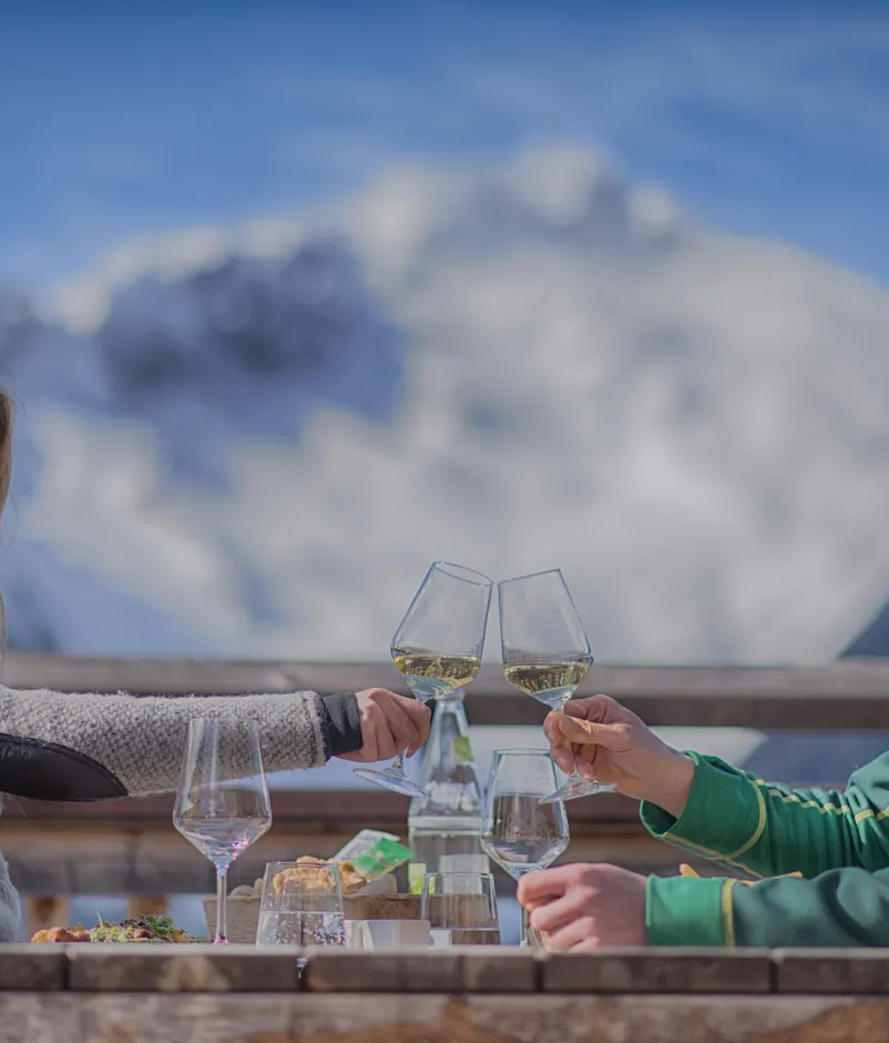 Paar stößt mit Weißwein auf Hüttenterrasse an, schneebedeckte Berge im Hintergrund