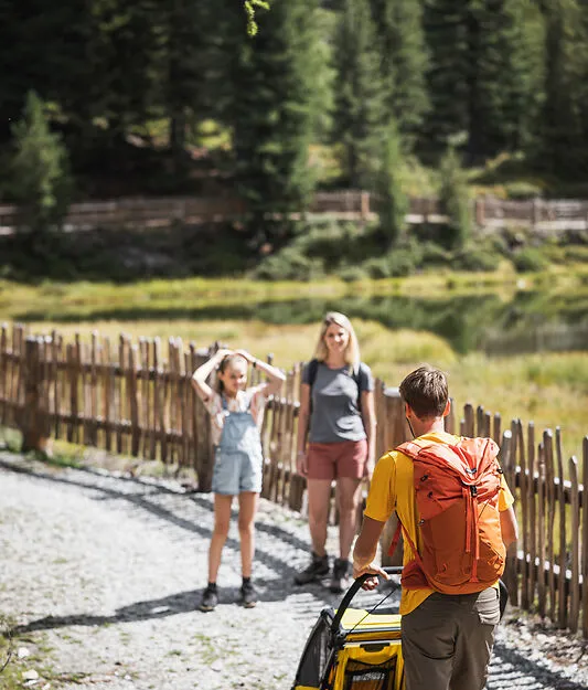 Familie mit Kinderanhänger auf Naturweg am Bergsee mit Holzzaun und Nadelwald im Ahrntal
