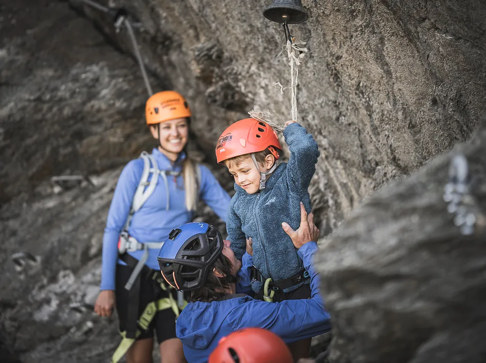 Familie am Klettersteig Ahrntal: Vater hebt Sohn mit Helm zur Gipfelglocke, Mutter freut sich