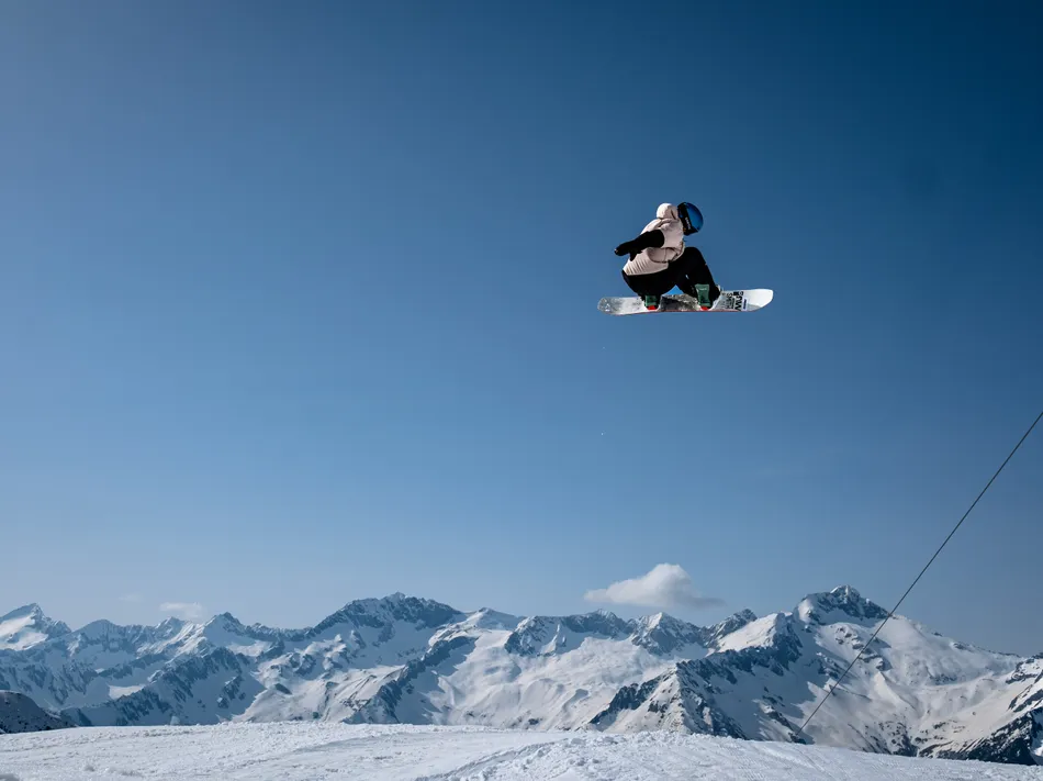Snowboarder beim hohen Sprung über den Snowpark-Kicker mit Seilbahn und Alpenpanorama