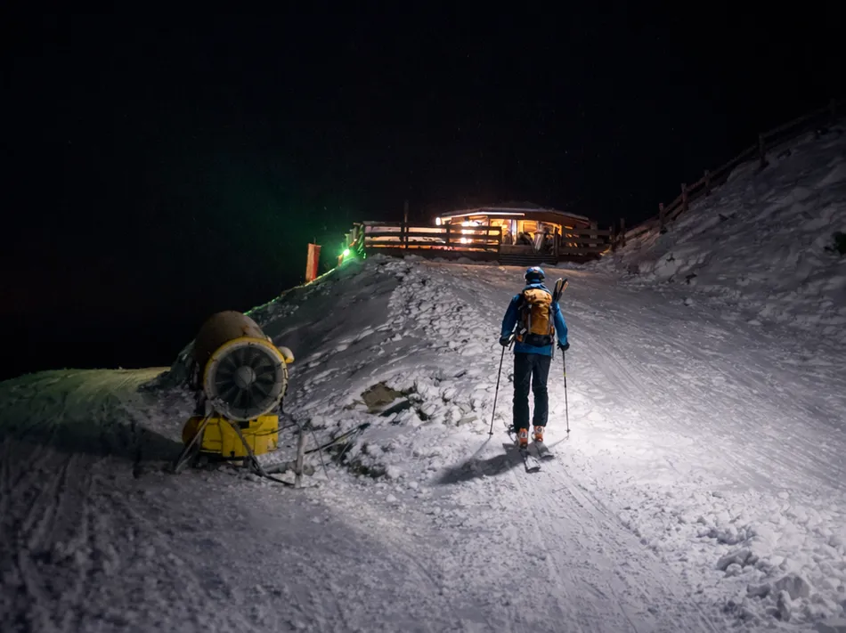 Skitourengeher mit Stirnlampe steigt nachts zur erleuchteten Berghütte am Klausberg auf