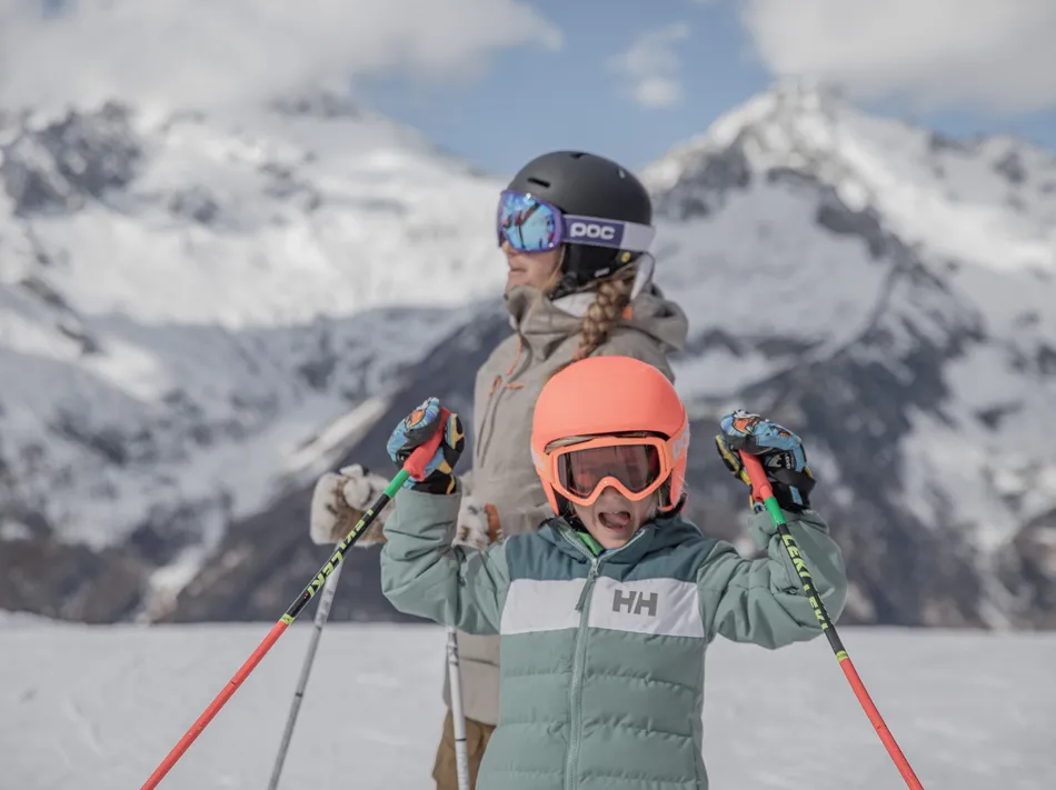 Jubelndes Kind mit orangem Skihelm hebt Stöcke auf der Piste, Mutter im Hintergrund