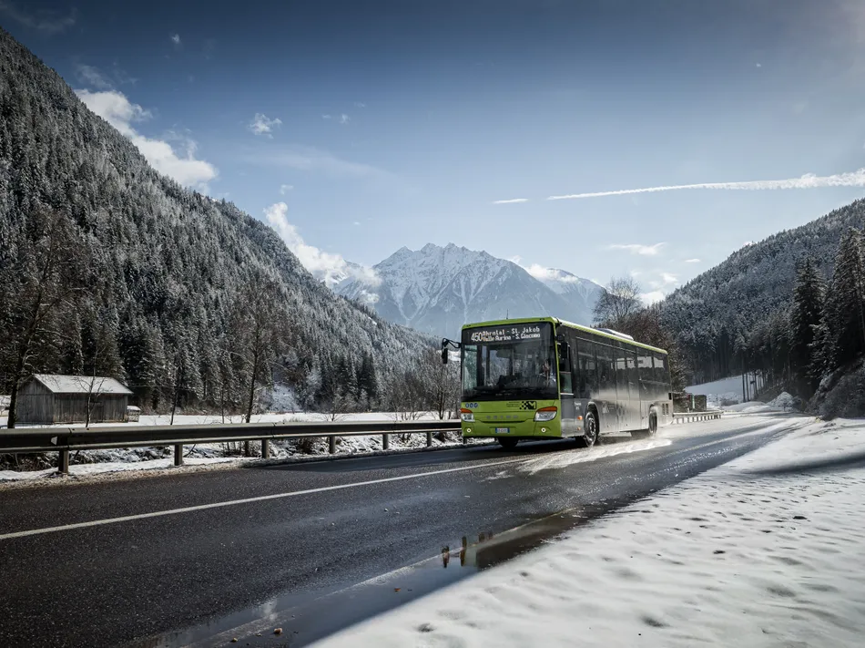 Green ski bus on Ahrntal line drives through snowy valley with forest and mountains